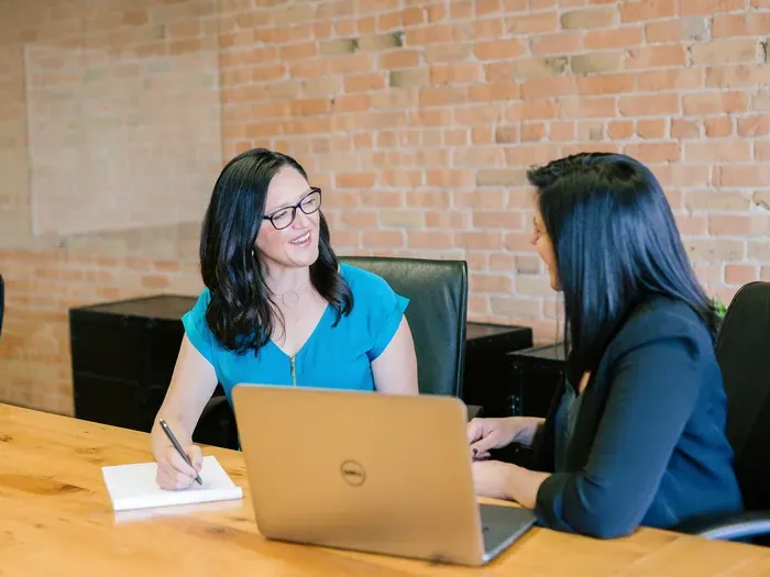 Two women meeting at a desk with a laptop and notes, discussing feedback and ratings for neighborhood services
